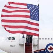 Mark Carney descends the steps of a plane hidden by an American flag. 
